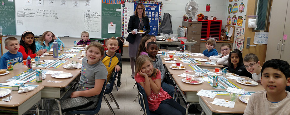 elementary students sit at a classroom table with plates and forks waiting for their Thanksgiving lunch