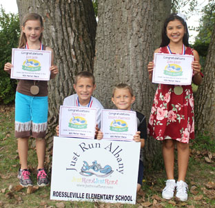 four elementary students stand near a tree showing off their running medals