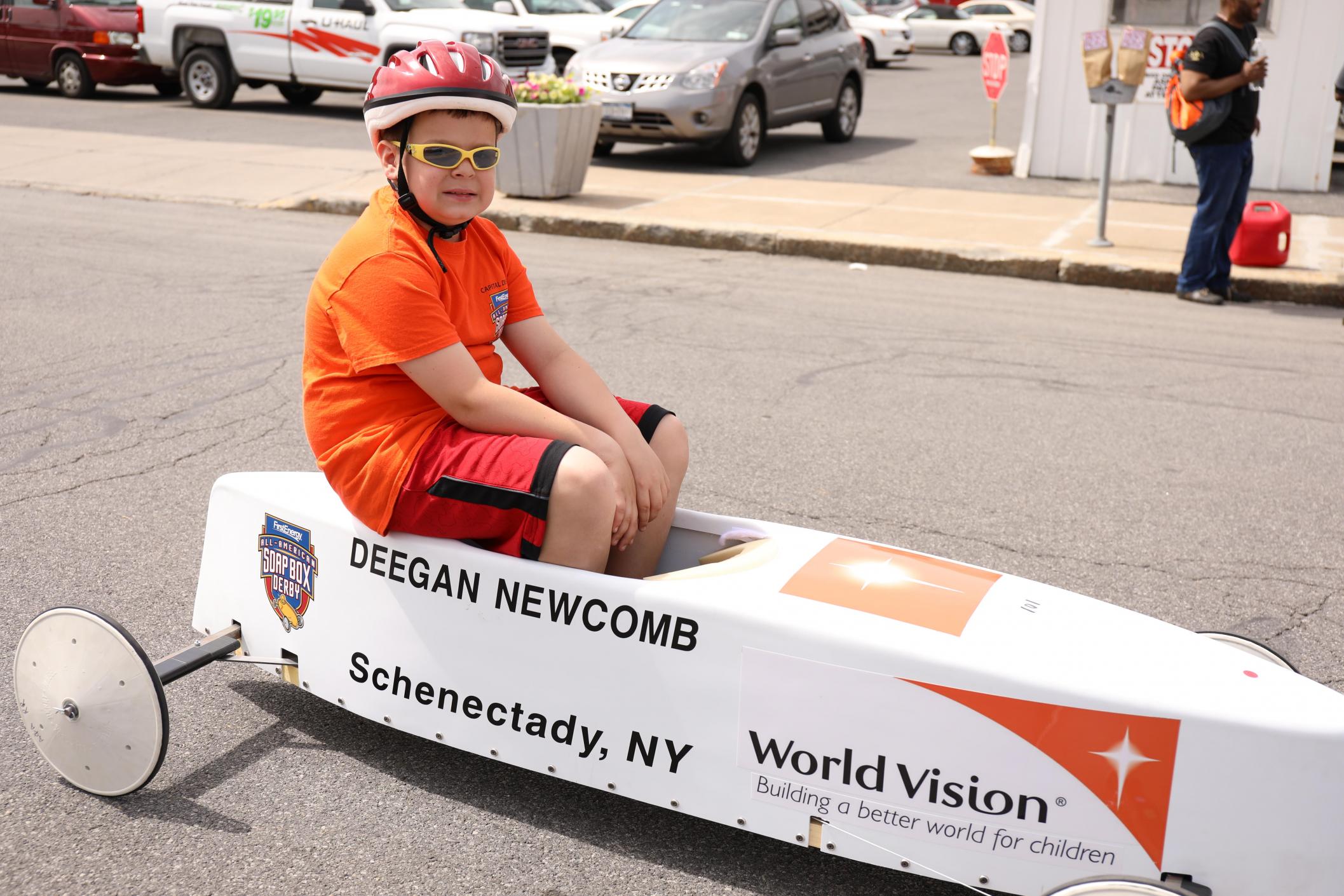 4th grader sits in his soap box derby car wearing a helmet and glasses