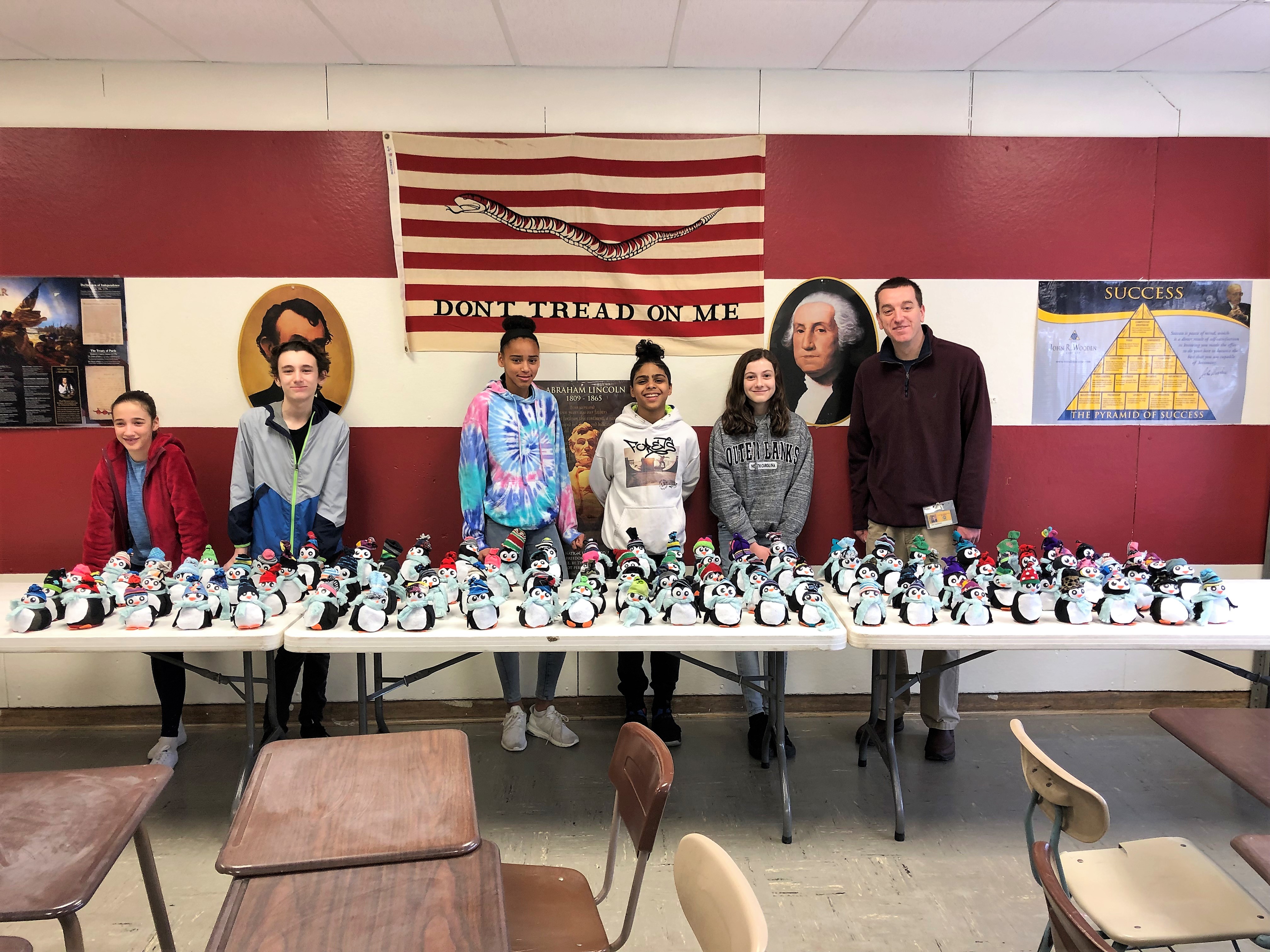 group of students in teacher standing behind a long table full of miniature stuffed penguins