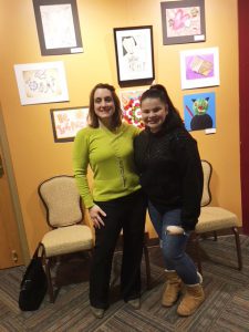 student stands with teacher in front of student artwork