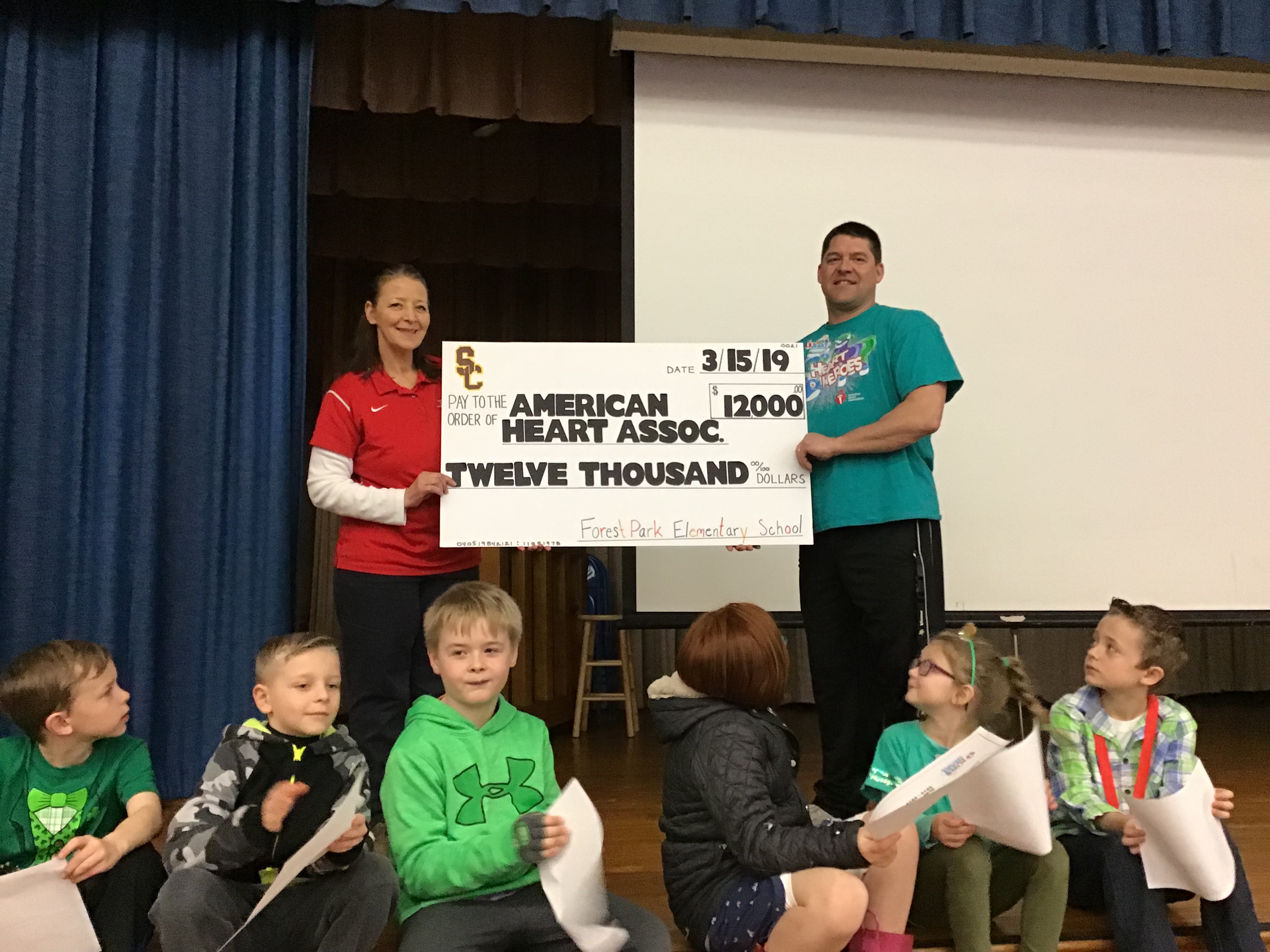 two teachers hold a big check while student winners sit on stage in front of them