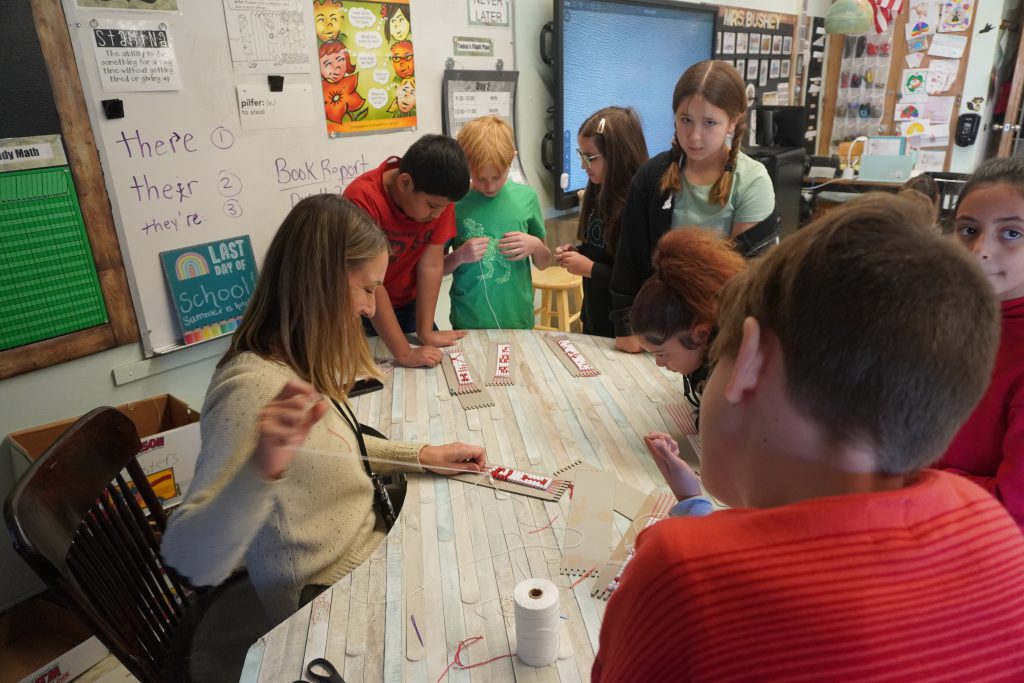 teacher is threading beads on a Wampum belt surrounded by students. 