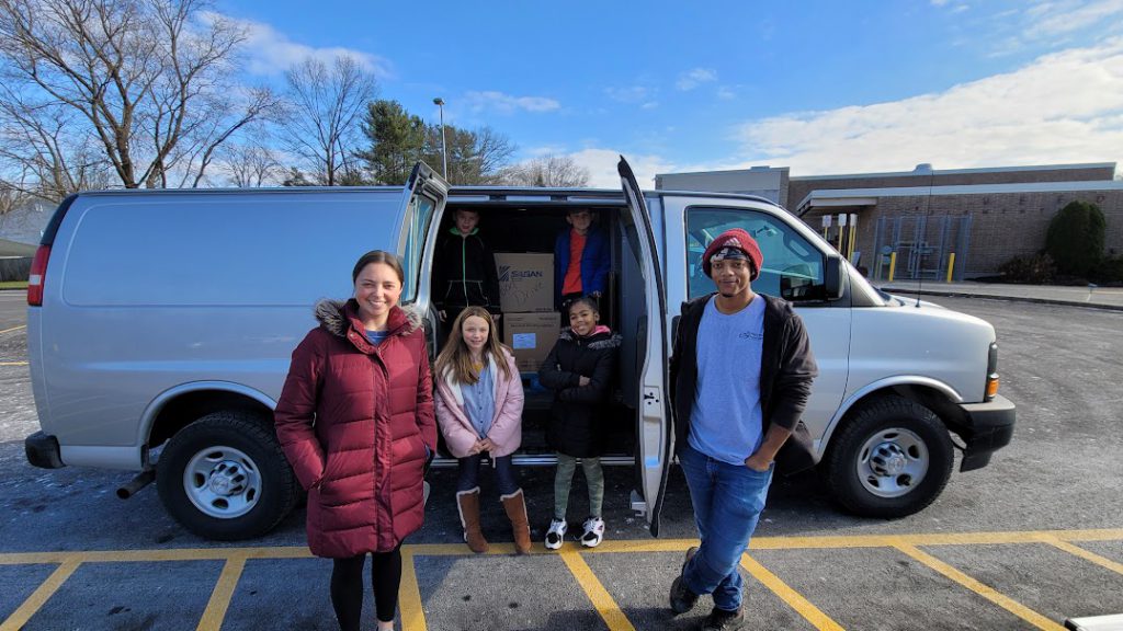 students and staff pose in front of a van full of boxes of food. 