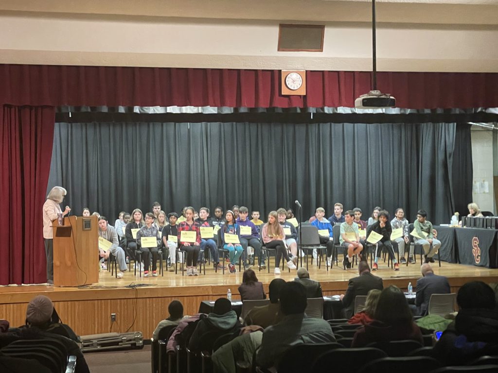 group of students sitting on stage while announcer talks into the microphone. 