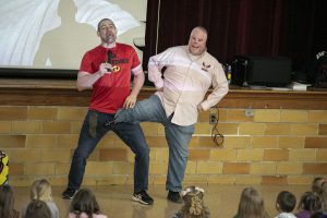 two guys standing together on a stage. one person has a burned face. 