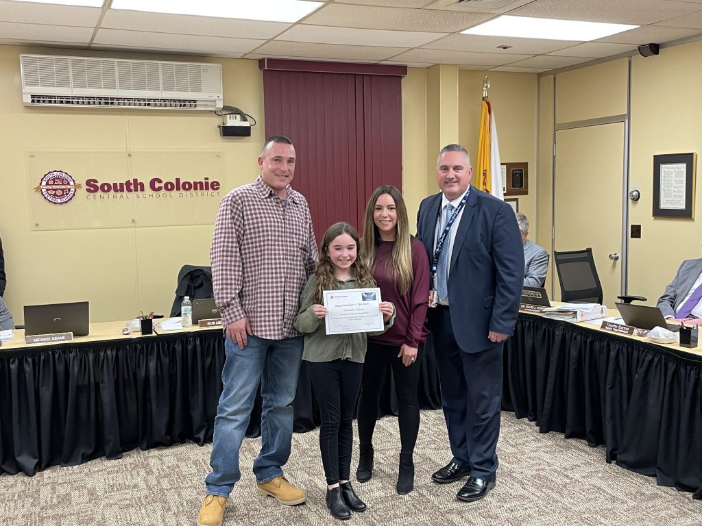 A mother, father and daughter pose with the Superintendent of Schools in the board room. The daughter is holding a certificate.