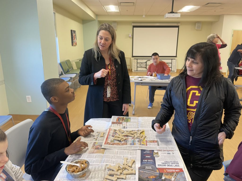 two adults interacting with studentds while they spread peanut butter on dog bones.