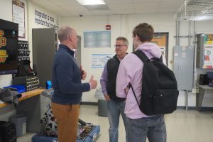 two adult males speaking with a student male inside a technology classroom.