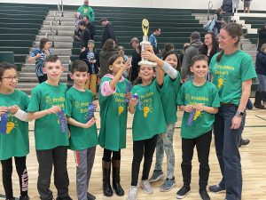 students stand with their teacher all wearing matching green tshirts and they hold their winning trophy up high.