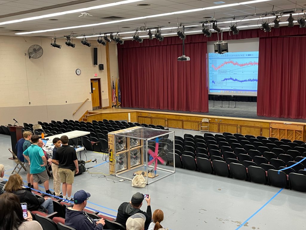 four kids stand next to a teacher and are getting ready to present their turbines at a conference