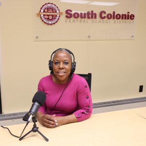 a female sitting in an office with a headset on in front of a microphone. The words South Colonie Central School District  hang behind her. 