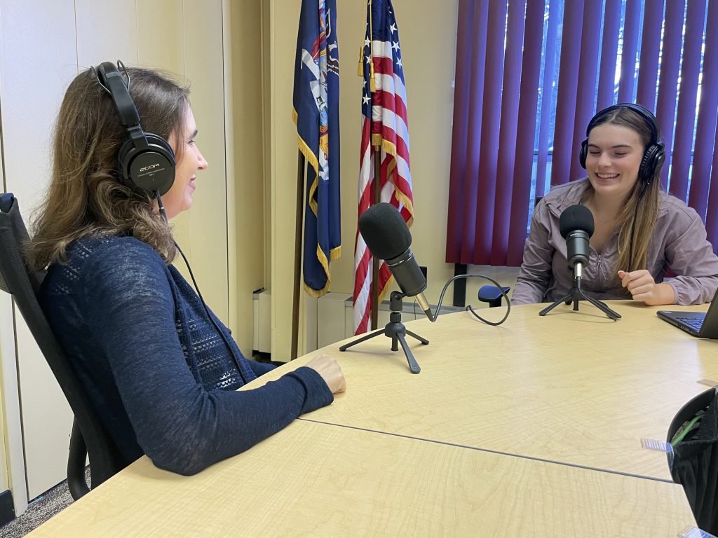 two women sitting in a room recording a podcast. two flags behind them. 