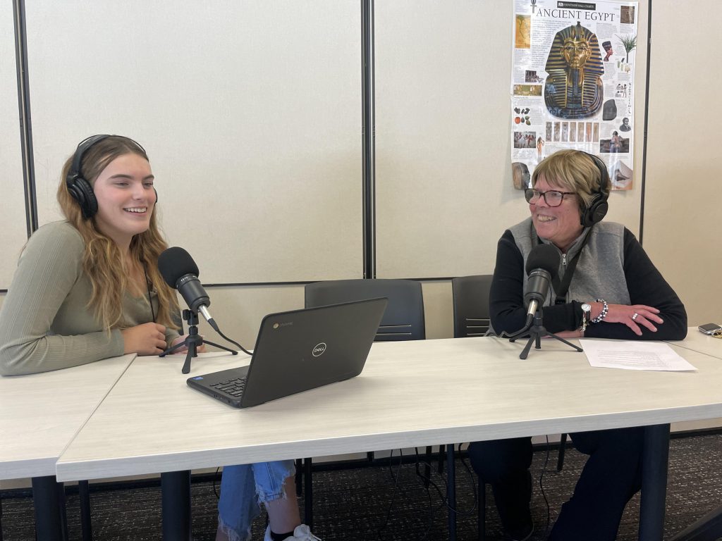 two people are sitting in an office space. a younger female is speakinginto a mic talking to an older female and they both have headphones on.