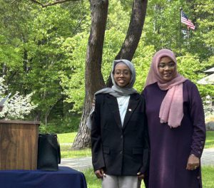 two females mother and daughter stand outside together posing for a photo. American flag is in the background.