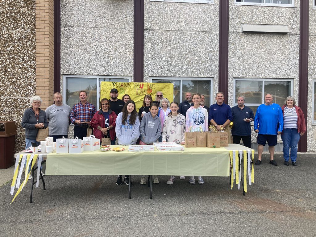 a group photo of students and staff infront of a school building with a sign behind them that says thank you bus drivers.