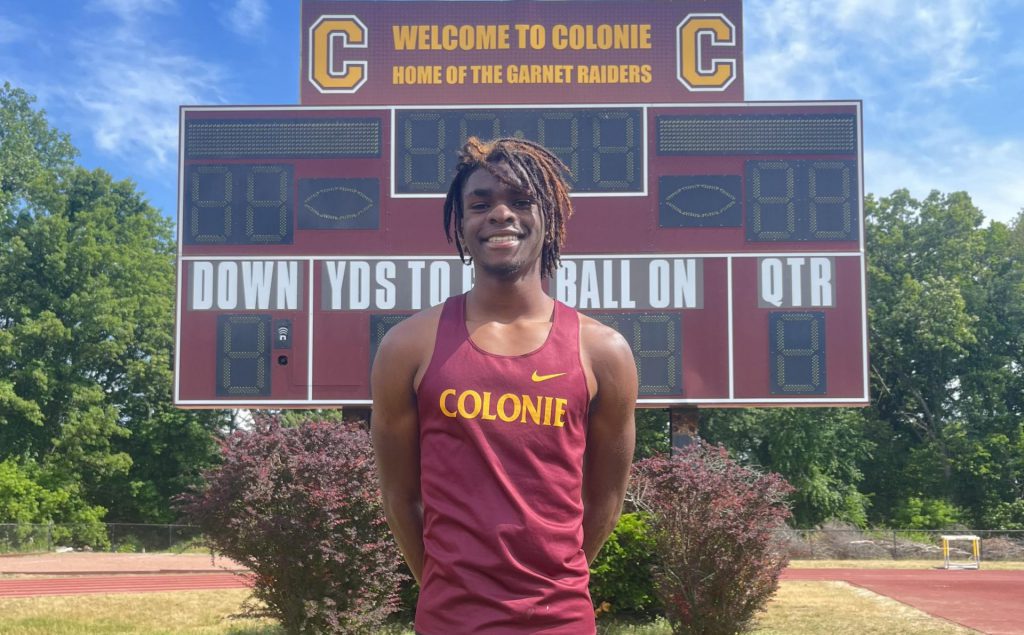 boys track athlete poses for a photo outside in front of a score board