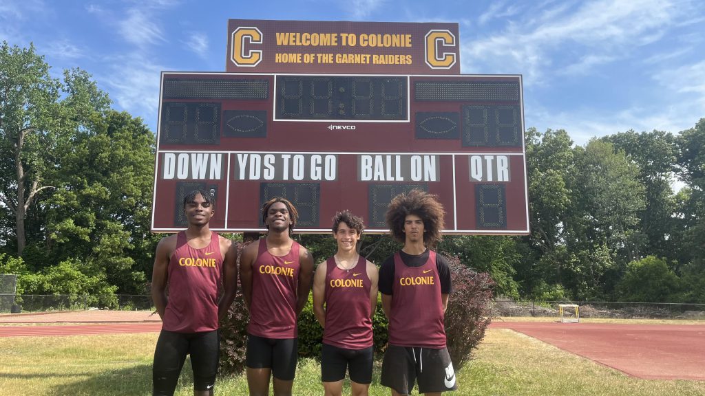 four track athletes standing posing for a photo in front of a Colonie scoreboard outside.