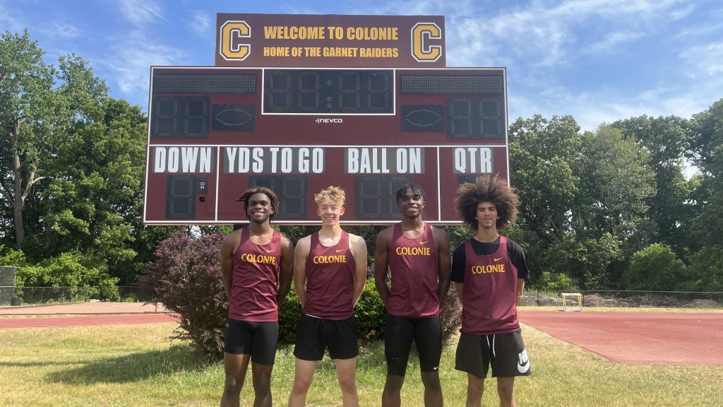 four track athletes standing posing for a photo in front of a Colonie scoreboard outside.