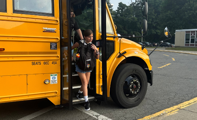 girl walks off the bus on the first day of school. Holds her backpack on the front of her body.