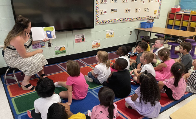 students sit on rug as teacher reads them a story.