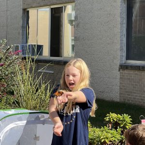 Child with monarch butterfly on their hand with wide-eyes and open mouth- clearly in shock and awe at the fantastic little creature who has chosen them as a perch.