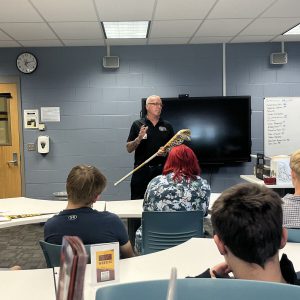 man speaks to students in a classroom setting. he is holding a lacrosse stick.