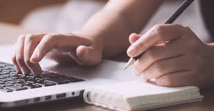 Right hand on keyboard of laptop, left hand holding pen over notepad.