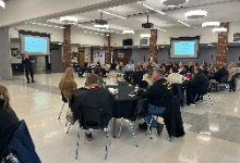 Man speaking in front of a crowd seated at tables in a cafeteria