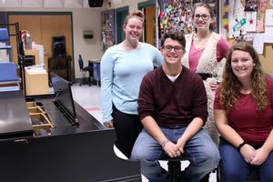 Two gilrs are standing and a boy and a girl are sitting at a piano